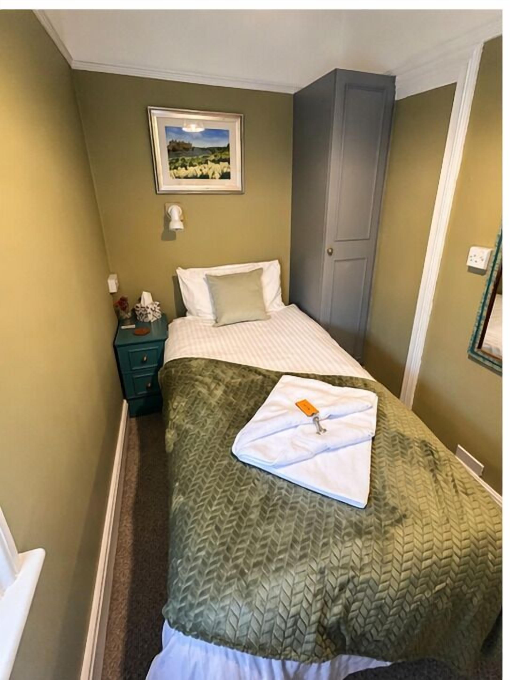 A narrow hotel room with a bed, a nightstand, a green wall, a framed picture, and a door.