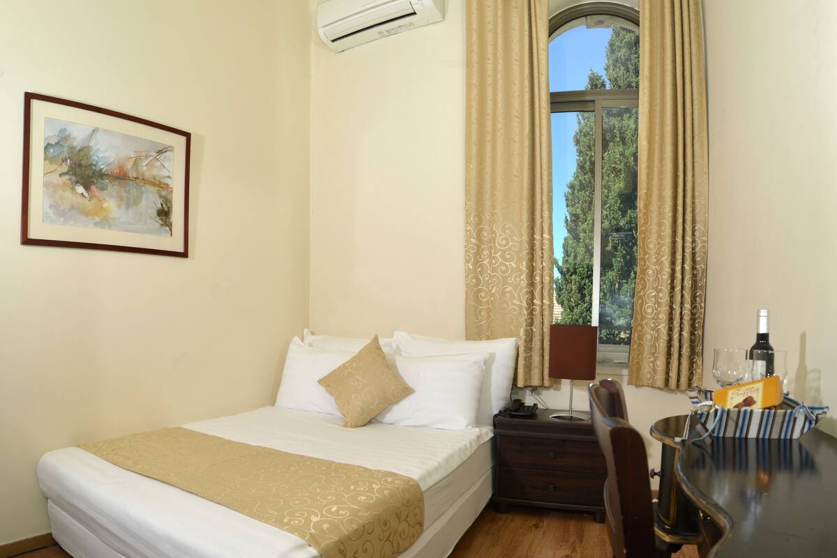 Quarto de hotel com uma cama, uma escrivaninha com cadeira, janela com cortinas, ar condicionado fixado na parede e um quadro emoldurado.