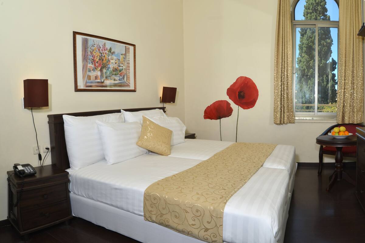 Quarto de hotel com uma cama grande, duas mesas de cabeceira, uma luminária, um quadro na parede, uma janela com cortinas e uma pequena mesa com frutas.