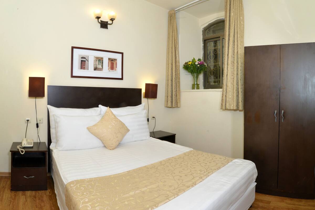 Cama bem arrumada com manta bege, duas luminárias de cabeceira, um quadro emoldurado sobre a cama, um guarda-roupa e uma janela com cortinas.
