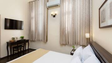 A hotel room with a bed, a desk with a chair, a TV mounted on the wall, an air conditioner, and curtains.