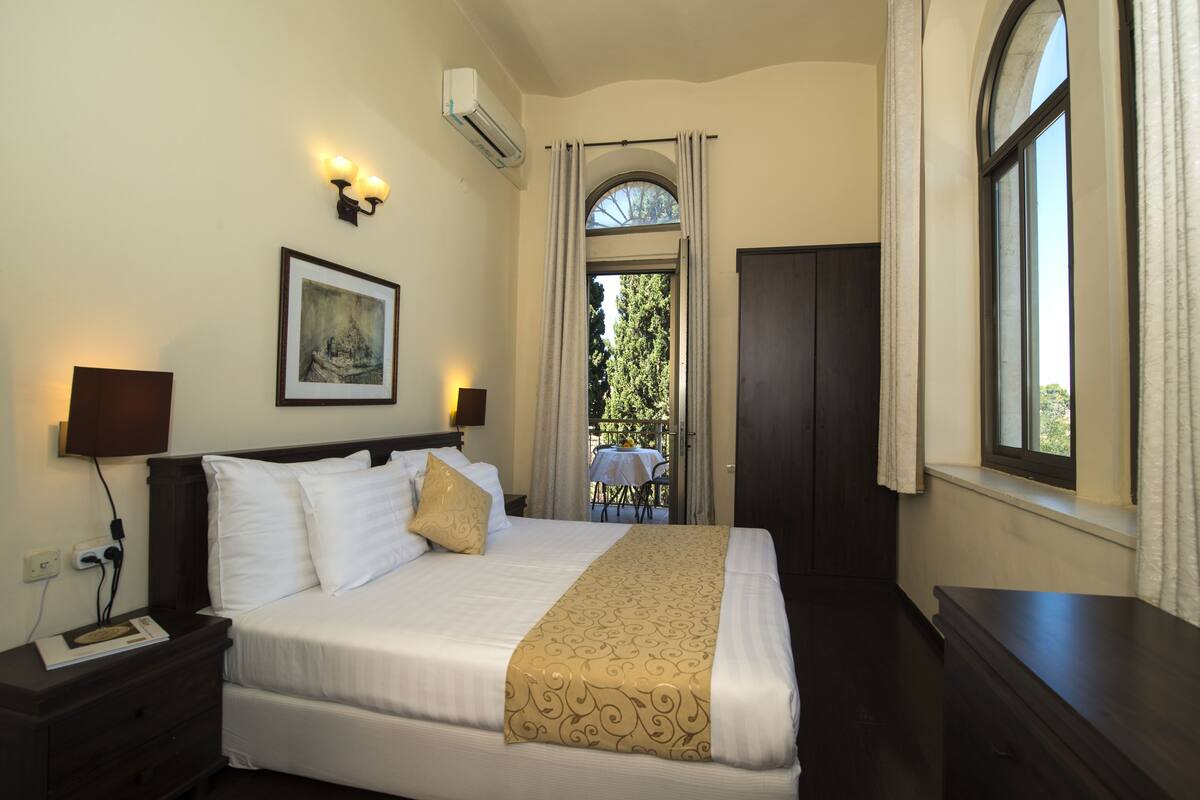Una cama bien tendida con sábanas blancas y un plaid estampado en dorado, una mesita de noche de madera oscura con una lámpara, un cuadro en la pared y una ventana con cortinas.