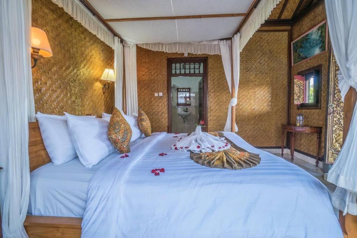 Una cama con ropa blanca, cabecera de madera y dosel. Hay una mesita con una lámpara y un marco de foto. La habitación tiene una pared con textura y una puerta que da al baño.