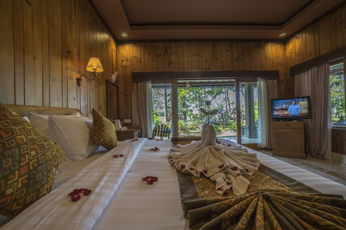 Habitación con paredes de madera, una cama con sábanas blancas, un televisor sobre un mueble de madera y vistas a zonas verdes al exterior.
