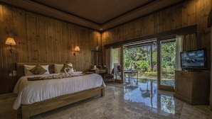 A spacious bedroom with a large bed, wooden walls, a TV, and a view of a garden through a glass door. - Cendana Resort & Spa (Ubud)