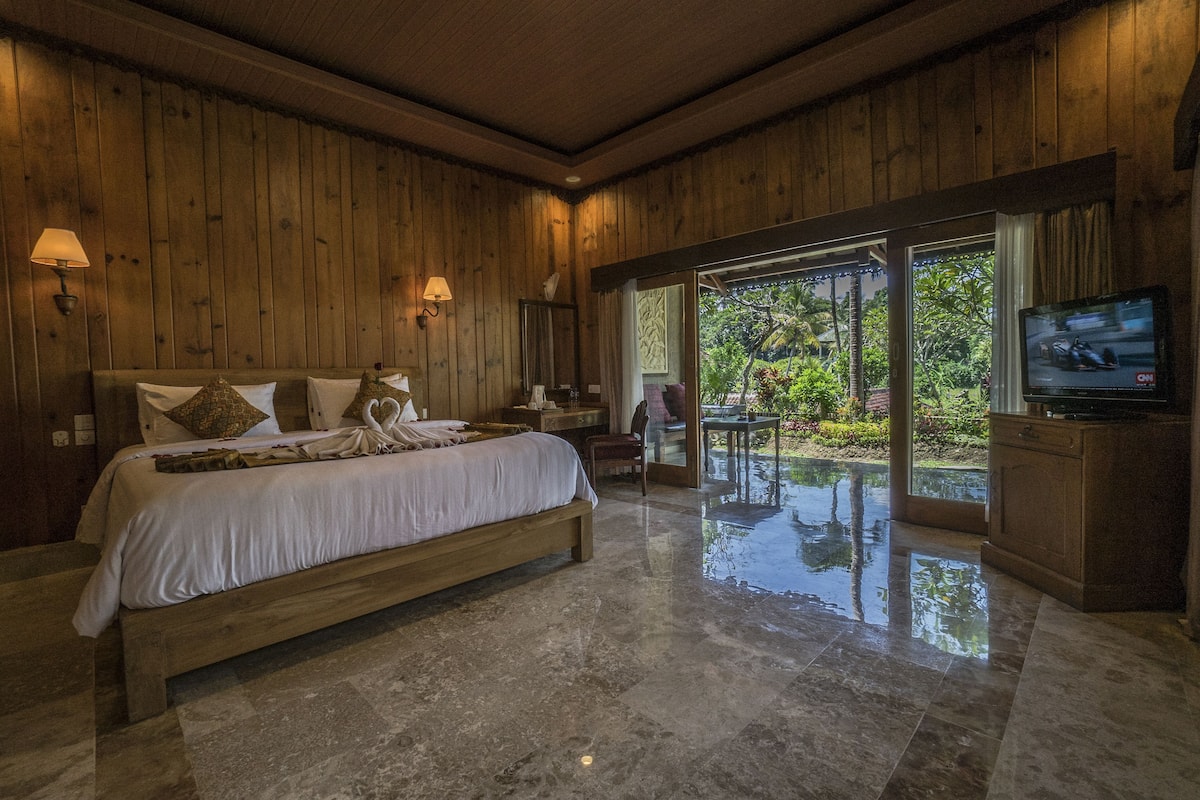 Un dormitorio espacioso con una cama grande, paredes de madera, un televisor y vistas de un jardín a través de un ventanal.