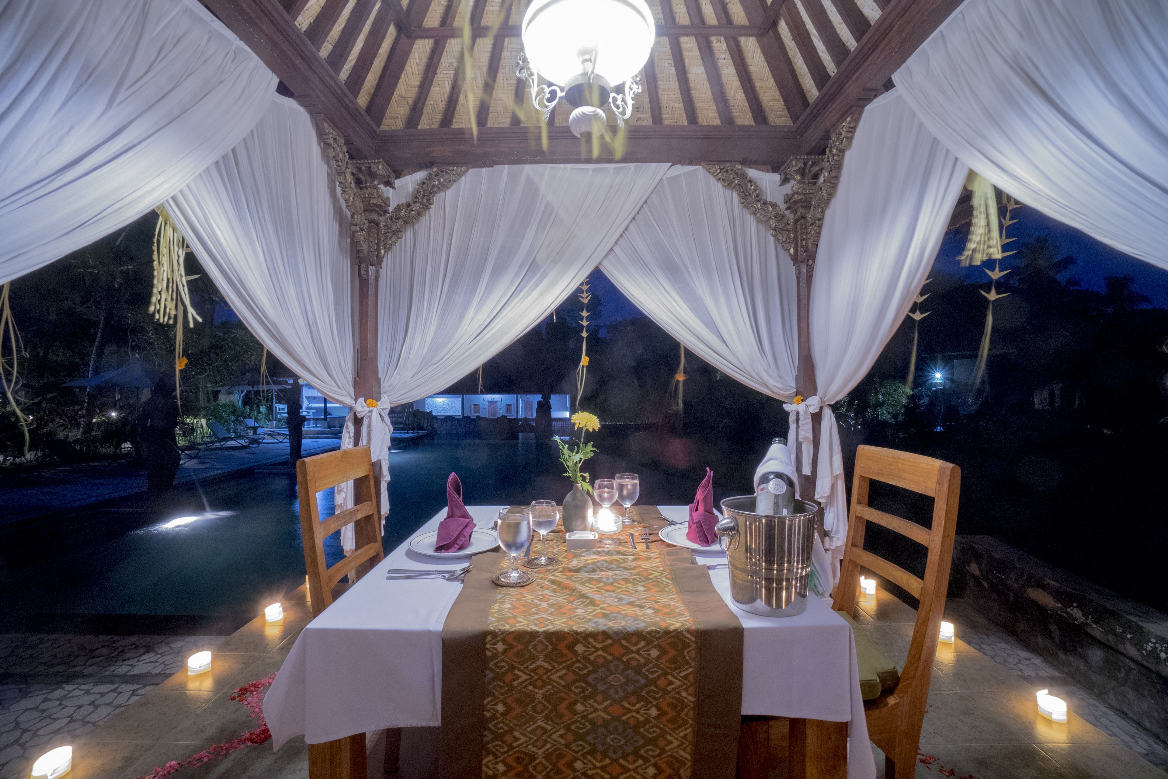 Un espacio de comedor al aire libre con un pabellón, cortinas blancas, iluminación con velas y una mesa dispuesta para dos.