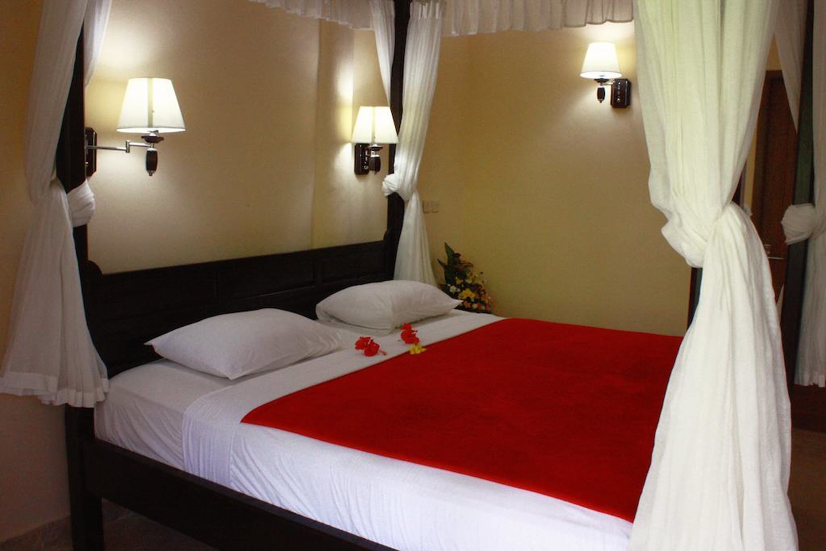 A bed with white linens, a red blanket, and white curtains. There are two wall-mounted lamps on either side of the bed.