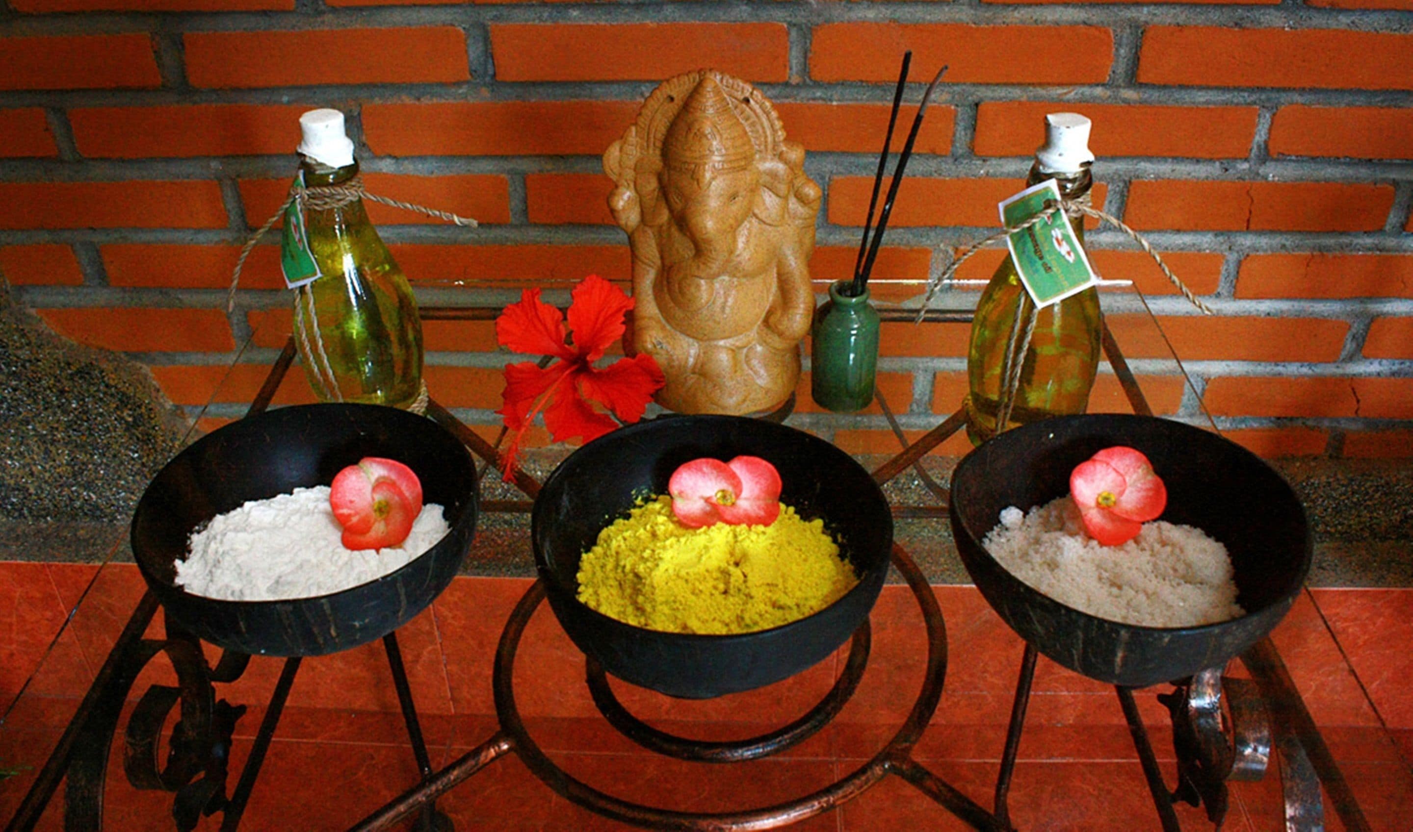 Three black bowls on a metal stand with white and yellow substances, possibly food, each with a red flower on top. Behind them are two green bottles with white caps, a decorative wooden figure, and a red flower on a brick wall.