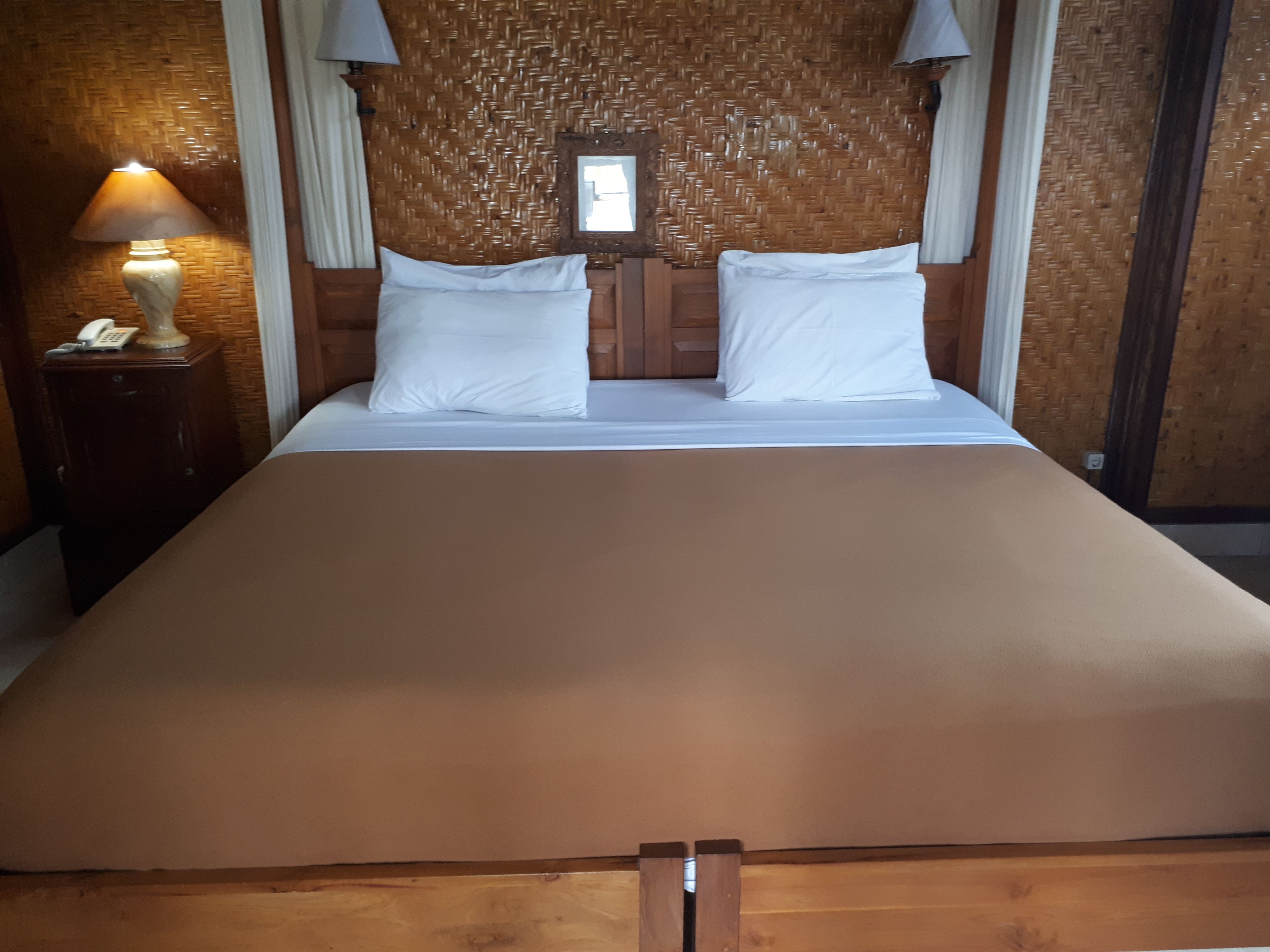 Un grande letto con lenzuola bianche, una testiera in legno e un comodino con lampada e telefono.