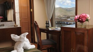 A hotel room with a bed, a wooden desk, a chair, a wooden wardrobe, a window with a view of mountains, and a vase with flowers. - Del Prado Inn (Cusco)