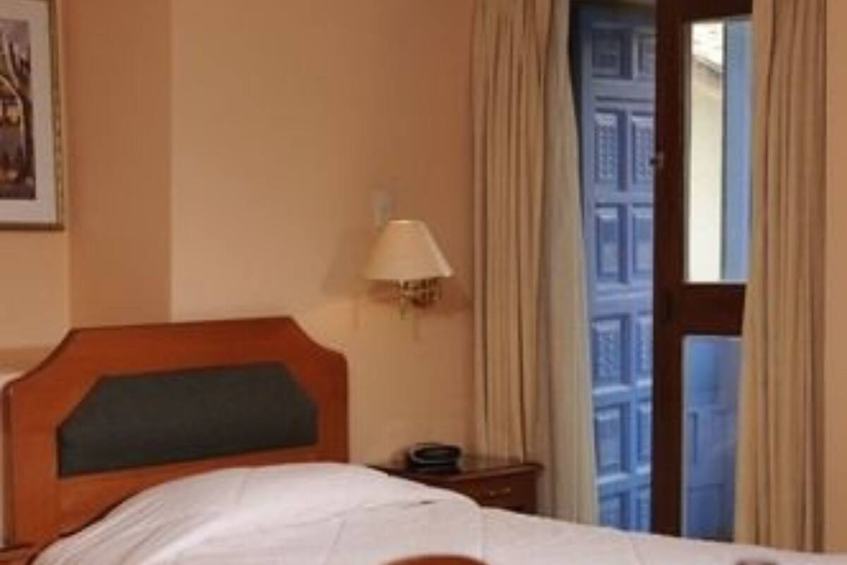 A hotel room with two beds, a table with wine glasses and a bottle, a lamp, and a window with curtains.