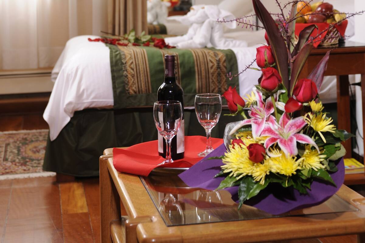 A hotel room with a bed, a table with a bottle and glasses, and a floral arrangement.