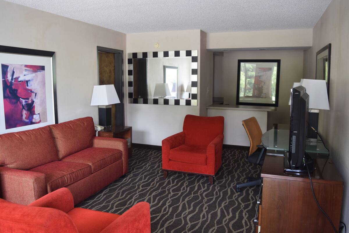 A hotel room with a red sofa, a red armchair, a desk with a computer, a lamp, and a mirror.