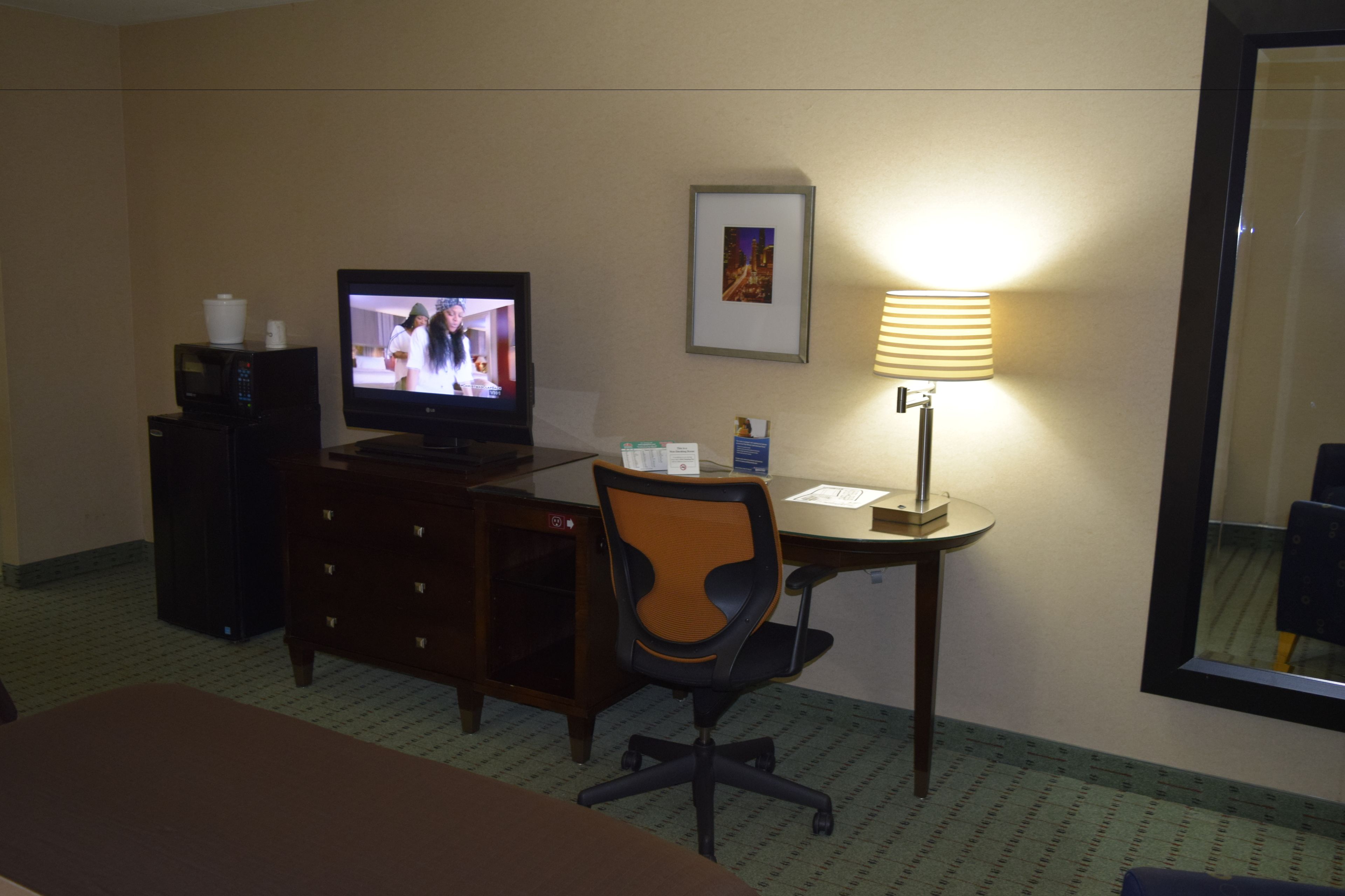 A hotel room with a television, desk, chair, lamp, and a mirror.