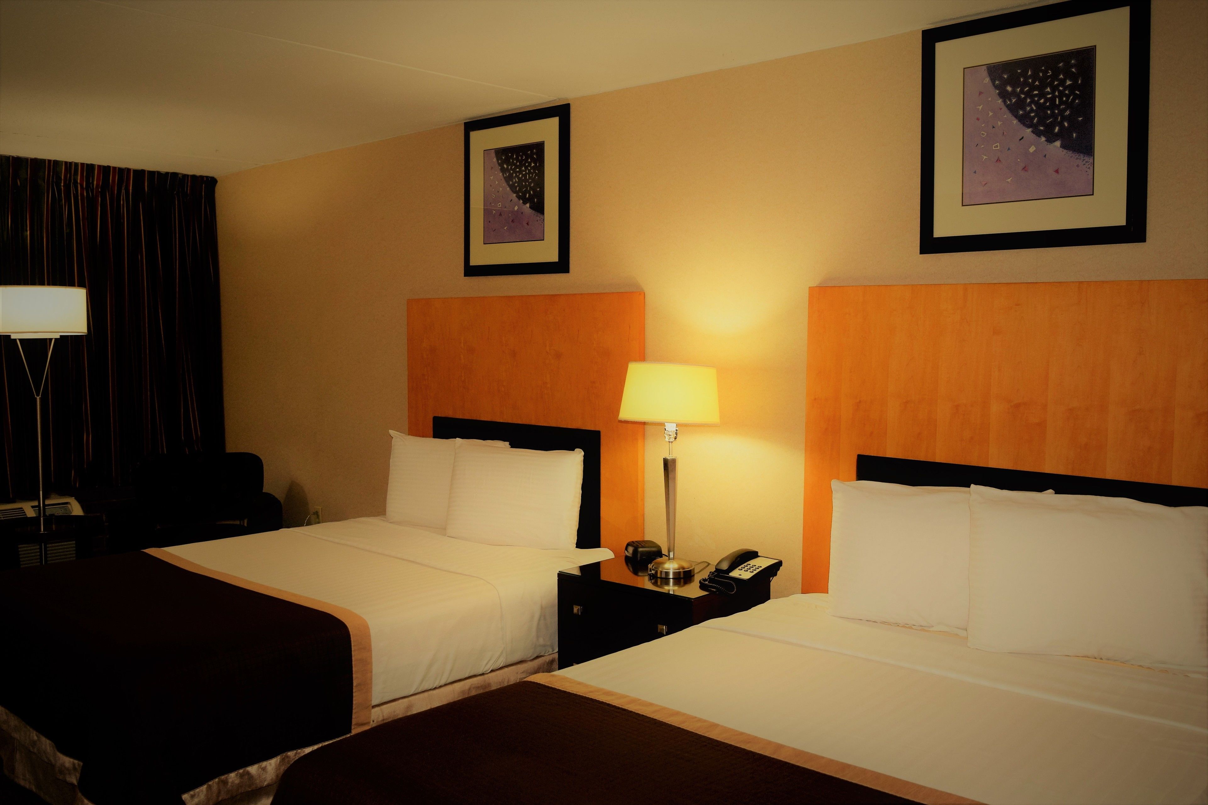 A hotel room with two beds, a nightstand with a lamp, a telephone, and two framed pictures on the wall.