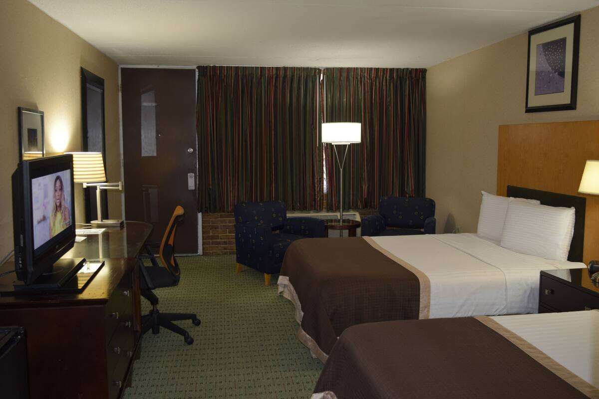 A hotel room with two beds, a desk with a computer, a chair, a lamp, and a television.