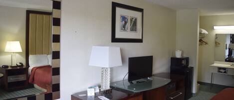 A hotel room with a bed, a desk with a computer, a TV, a mirror, and a bedside table with a lamp.