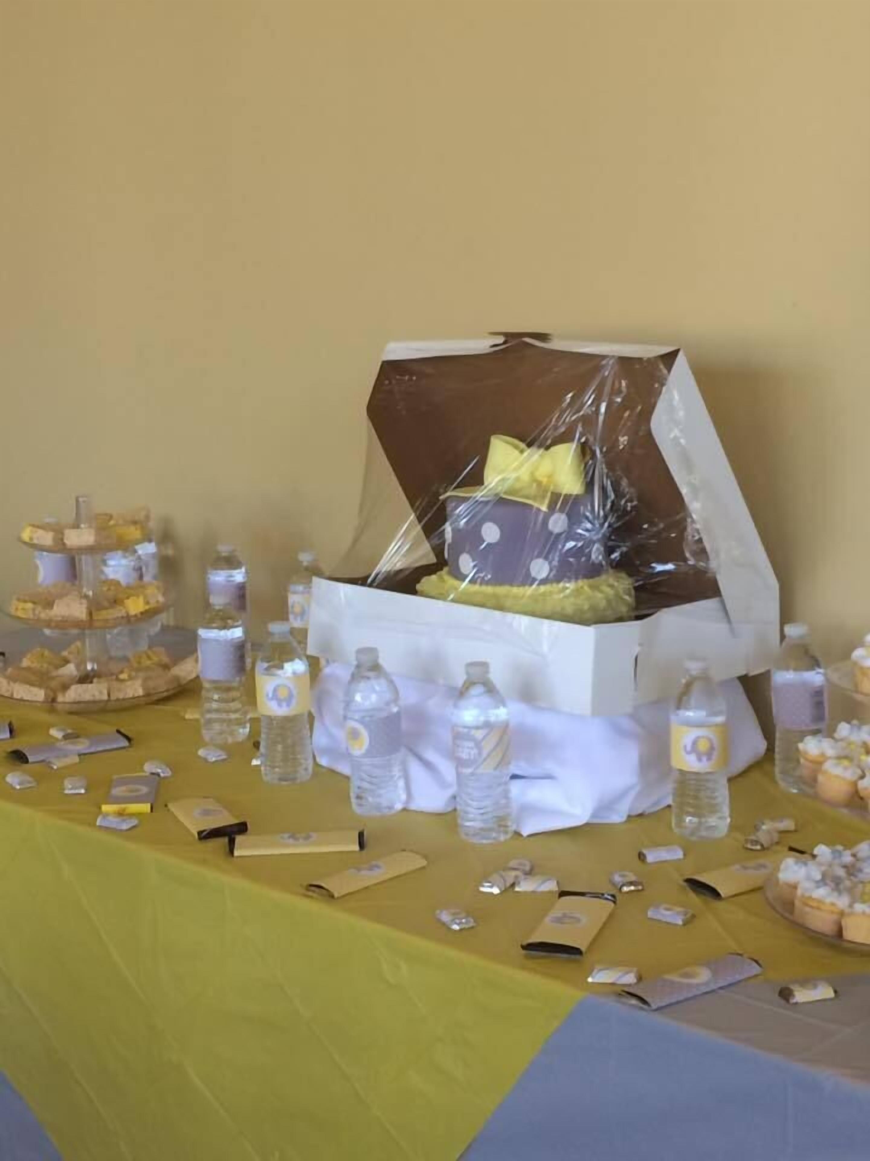 A table with a yellow tablecloth, a cake in a white box with a yellow bow, and various snacks and drinks.