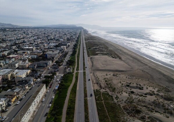 An aerial view of a coastal city with a long road running parallel to the beach, lined with buildings and vehicles.