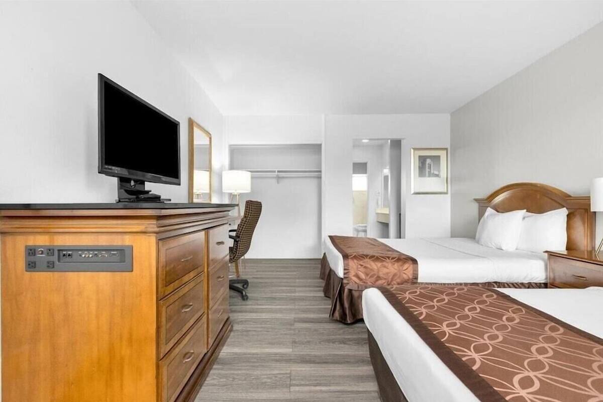 Habitación de hotel con cómoda de madera, televisor, cama con colcha marrón, escritorio con silla y mesita de noche con lámpara.
