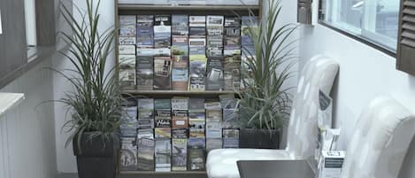 A reception area with a bulletin board displaying various brochures, two potted plants, a white upholstered bench, and a window with blinds.