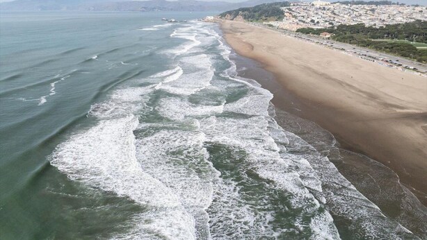 A beach with waves crashing onto the shore, a clear sky, and a distant cityscape. - Motel 6 San Francisco, CA – Great Highway (San Francisco)