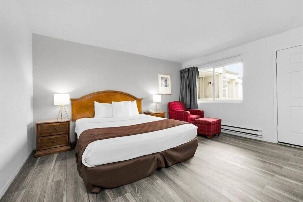 Una habitación de hotel con una cama grande, una mesita de noche de madera, un sillón rojo y una ventana con cortinas.