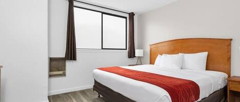 Quarto de hotel com uma cama de solteiro, uma janela com cortinas, uma mesa de cabeceira com abajur e cabeceira de madeira.