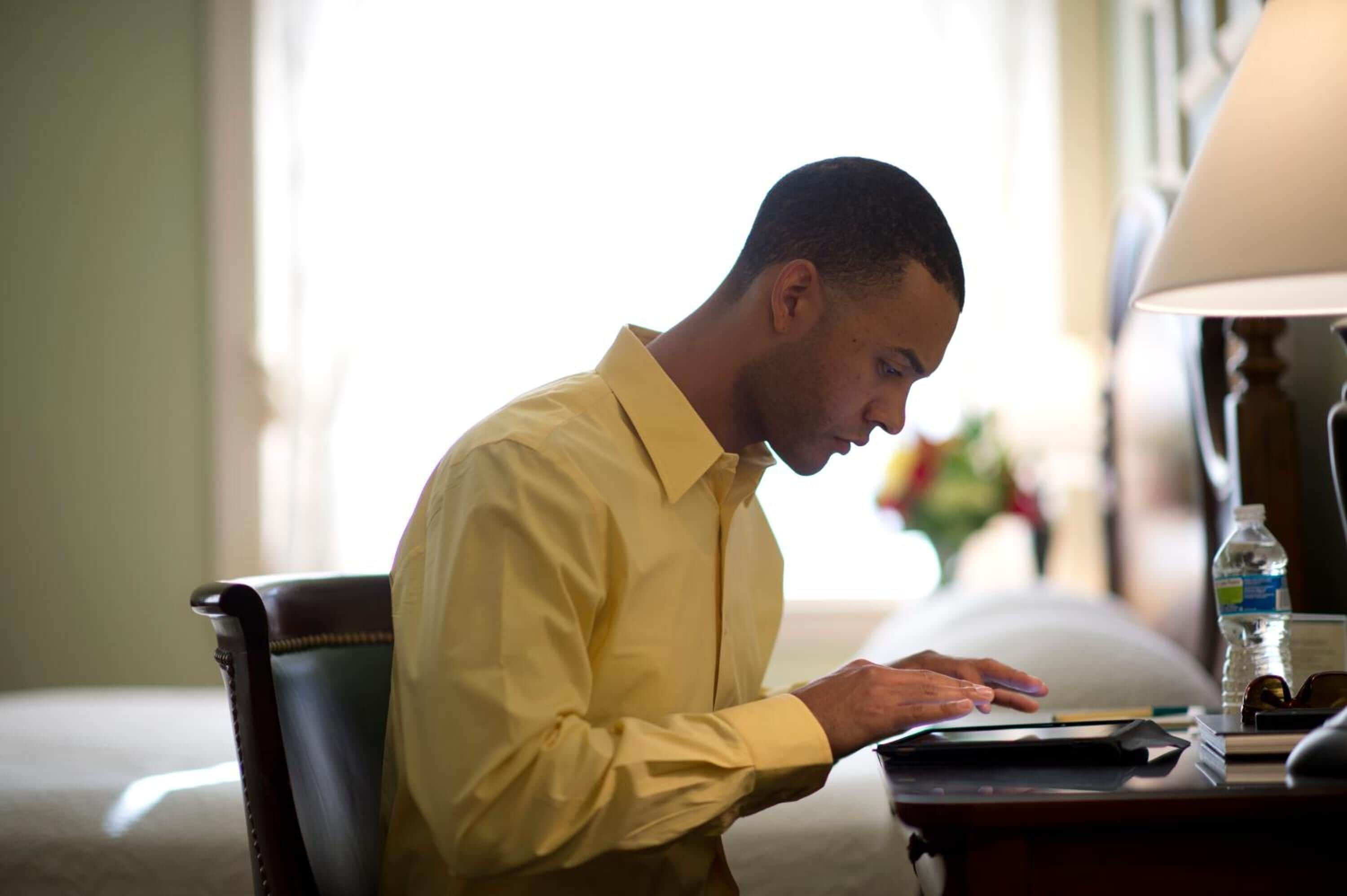 A man in a yellow shirt sitting at a desk with a laptop, a water bottle, and a lamp.
