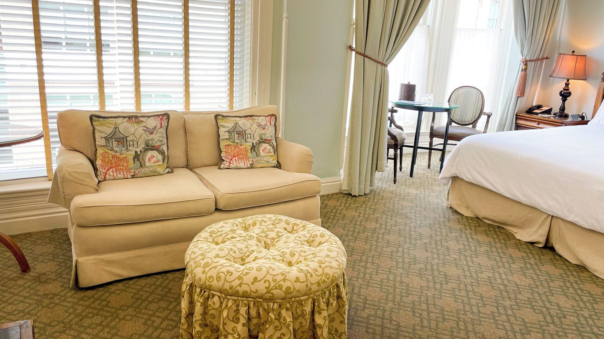 Quarto de hotel com sofá bege, duas almofadas decorativas, um banquinho redondo, uma pequena mesa redonda com uma cadeira, uma cama e uma mesa de cabeceira com abajur.