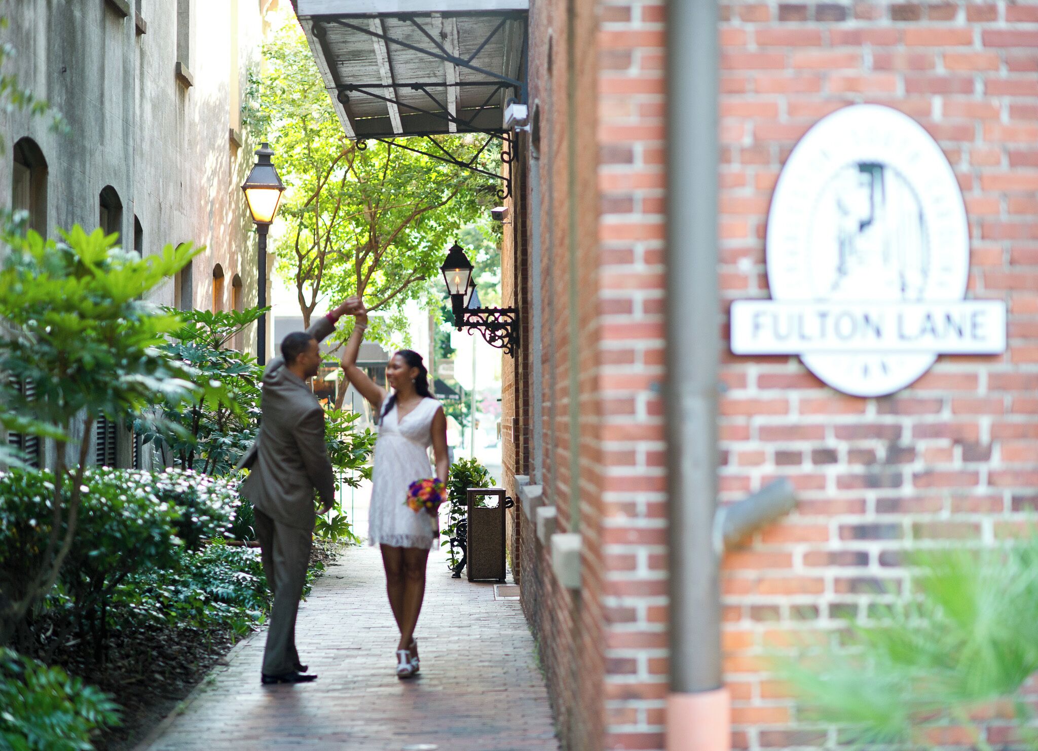 A couple walking down Fulton Lane, surrounded by greenery and brick buildings.