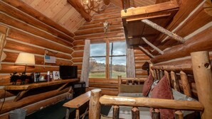 A log cabin interior with a wooden ceiling, a mounted deer skull, a television, a desk, and a window offering a view of the outdoors. - Hatchet Resort (Moran)