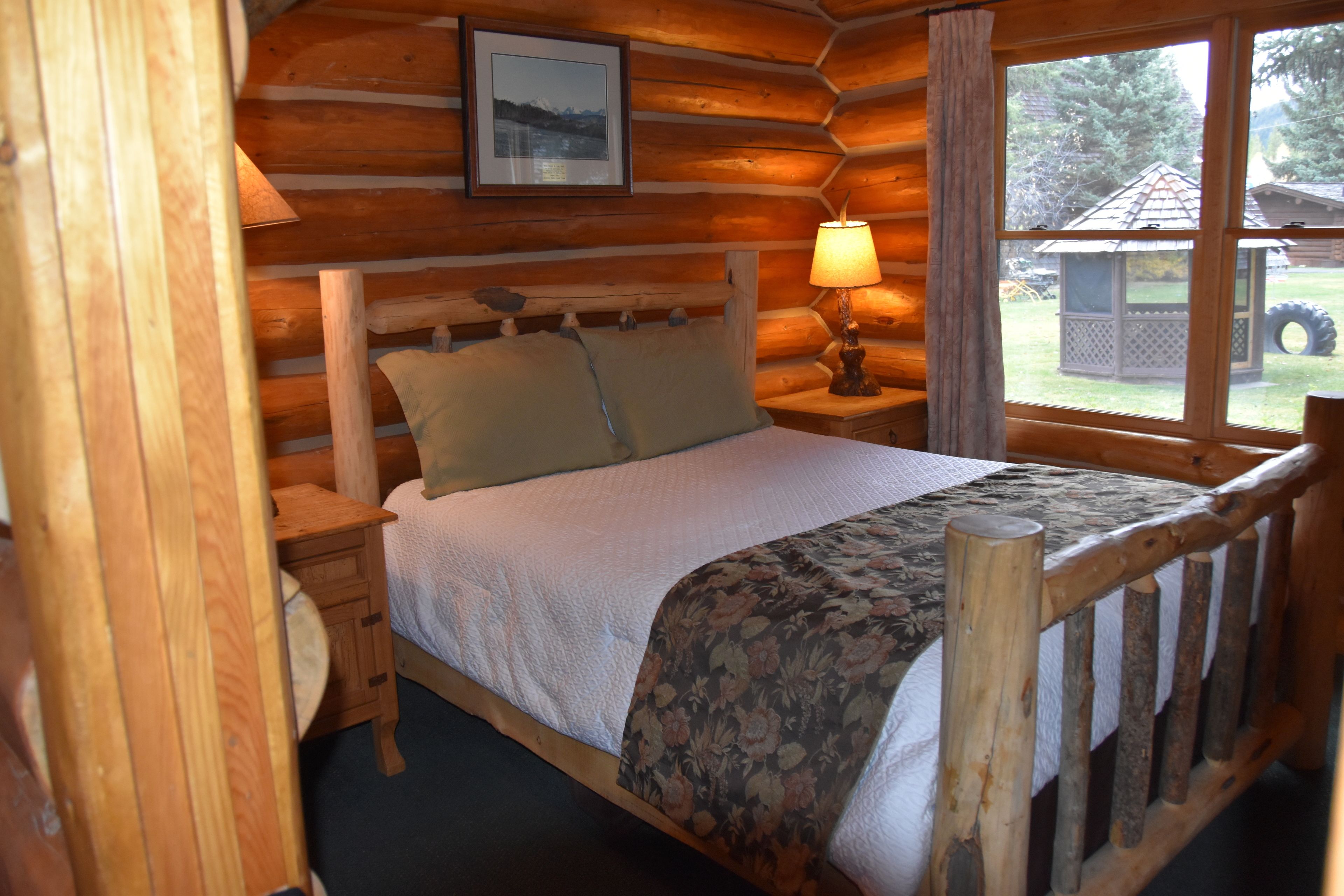 A wooden cabin bedroom with a bed, nightstand, lamp, and a window with a view of the outdoors.