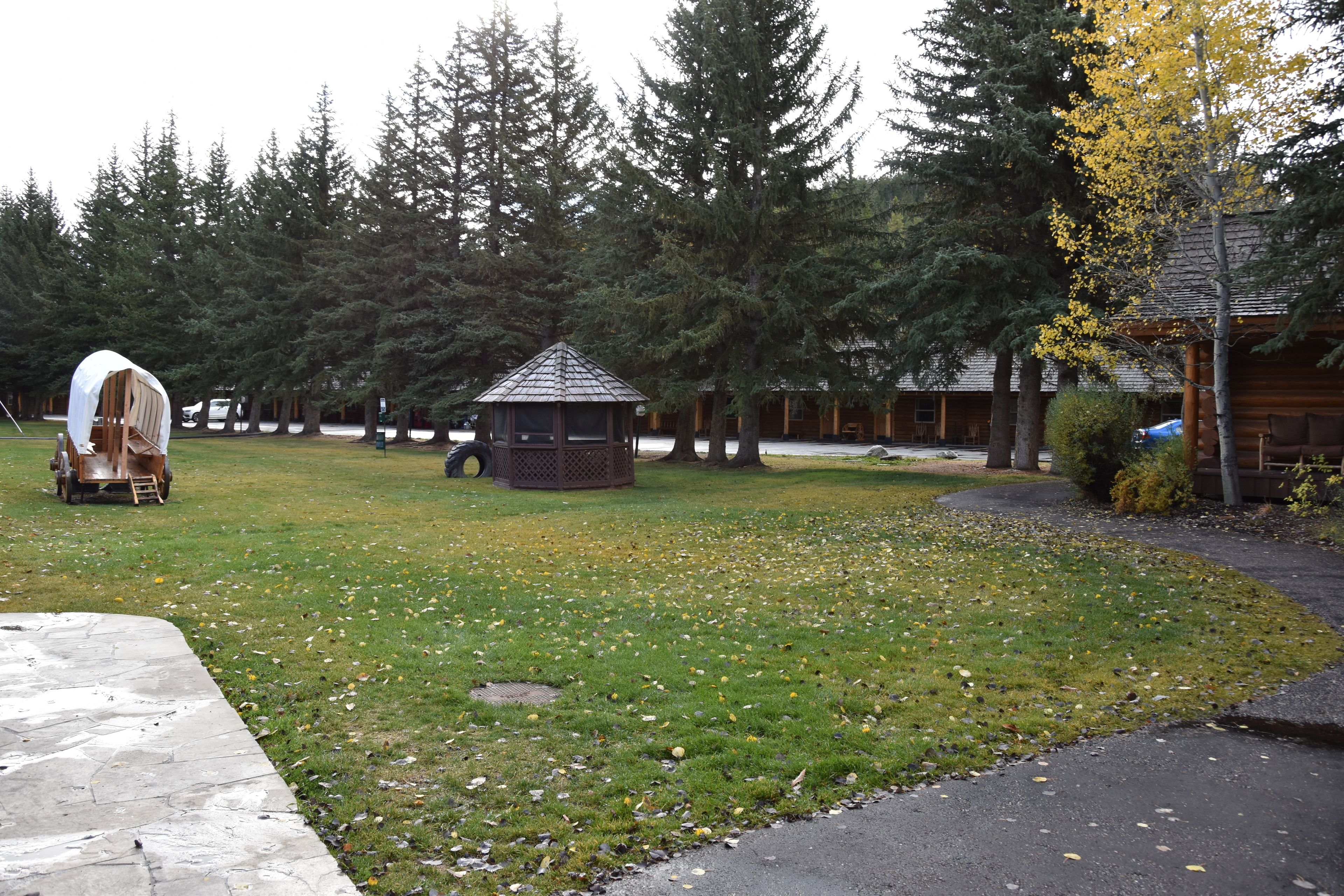 Un kiosque dans un parc, avec une charrette couverte et une cabane en rondins en arrière-plan.