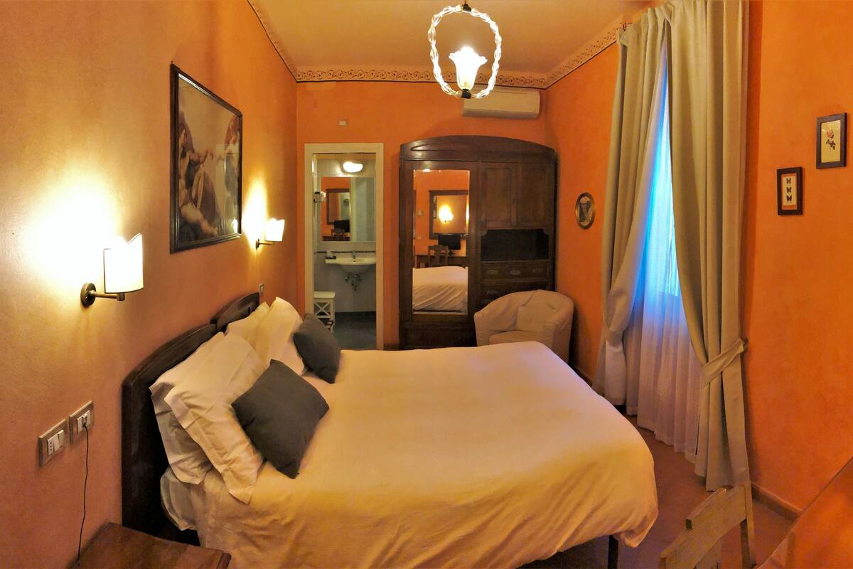 A hotel room with a bed, a bedside table, a mirror, a wardrobe, and a bathroom visible through an open door.