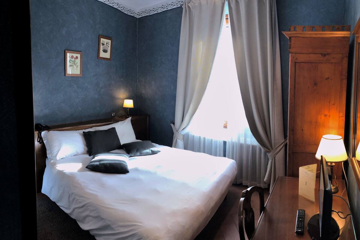A hotel room with a bed, a bedside lamp, a nightstand, a mirror, and a window with curtains.