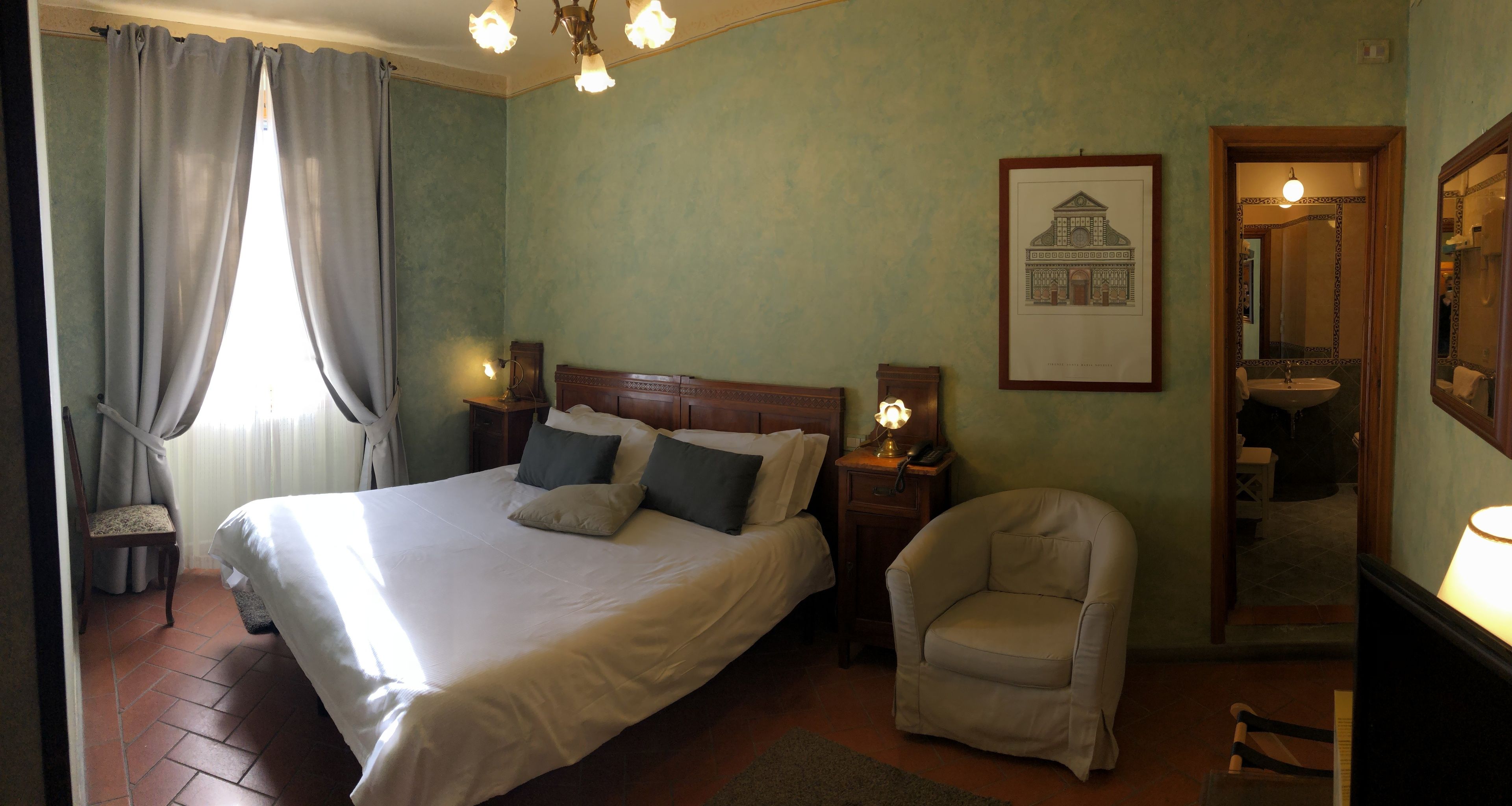 A bedroom with a large bed, a chair, a nightstand, a framed picture on the wall, and a bathroom visible through an open door.
