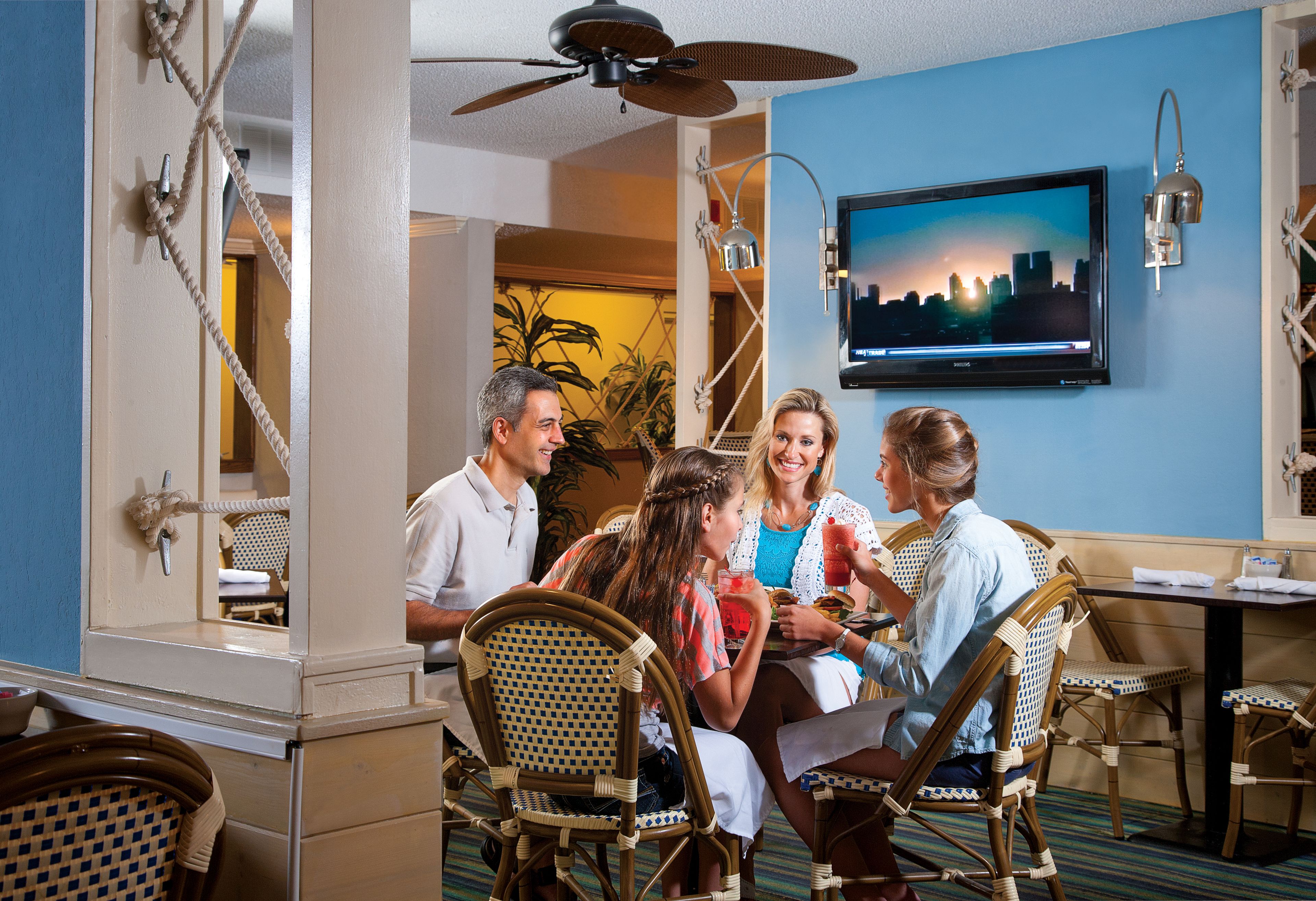 A hotel room with a cityscape TV, wicker chairs, a ceiling fan, and a dining table.