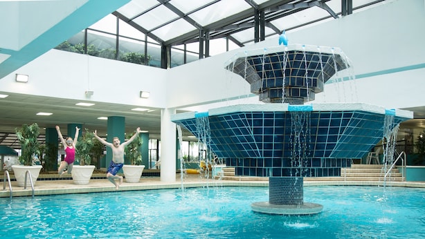 Una piscina interior con una fuente central, rodeada de plantas en macetas y un techo alto con tragaluces.