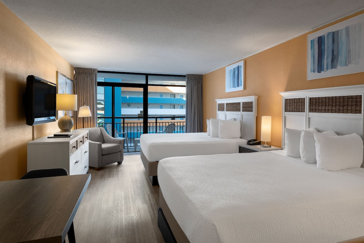 Habitación de hotel con dos camas, televisión, escritorio y balcón con vistas a edificios.