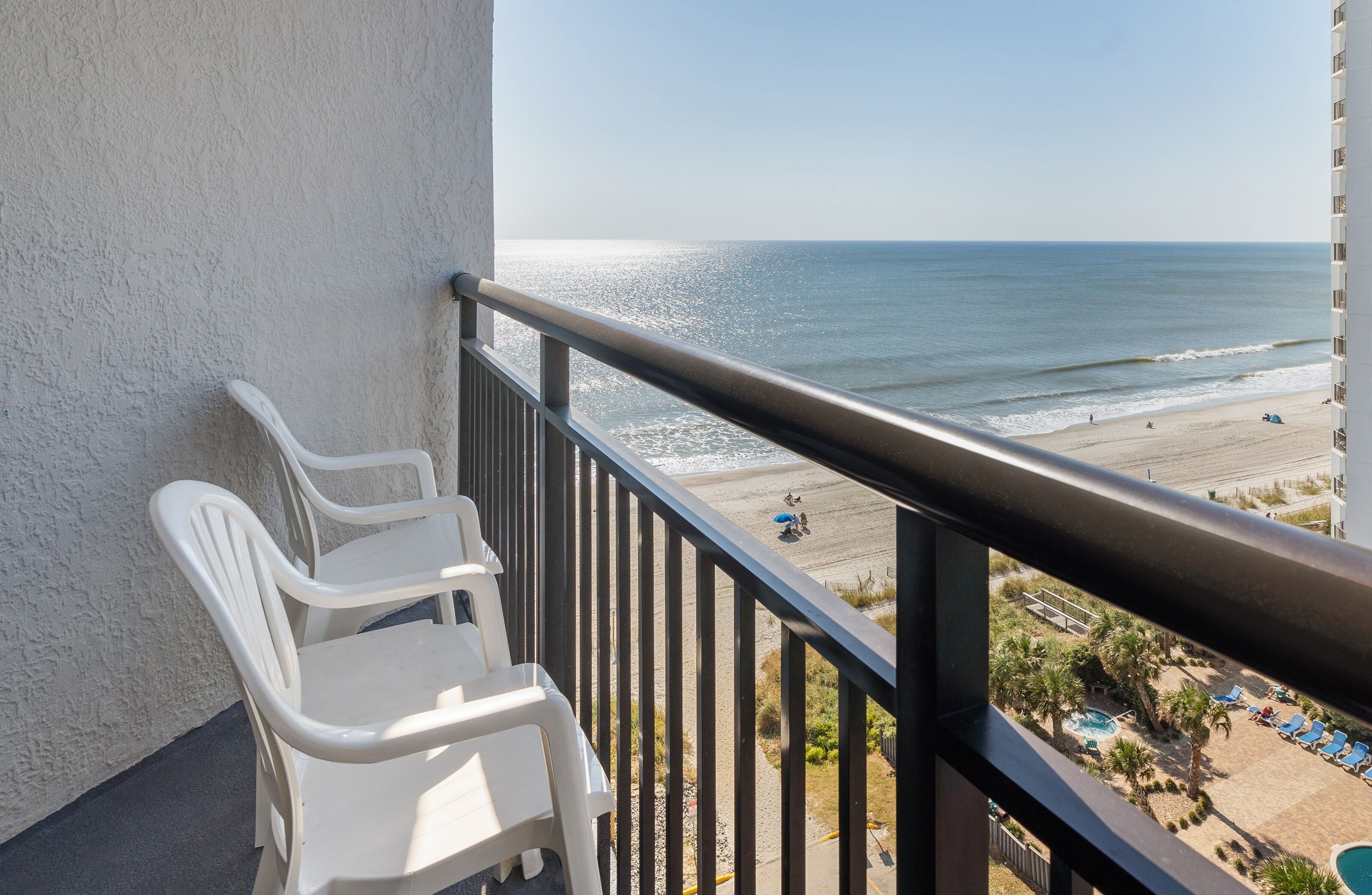 A balcony with white plastic chairs, a view of the beach, and a pool area with lounge chairs.