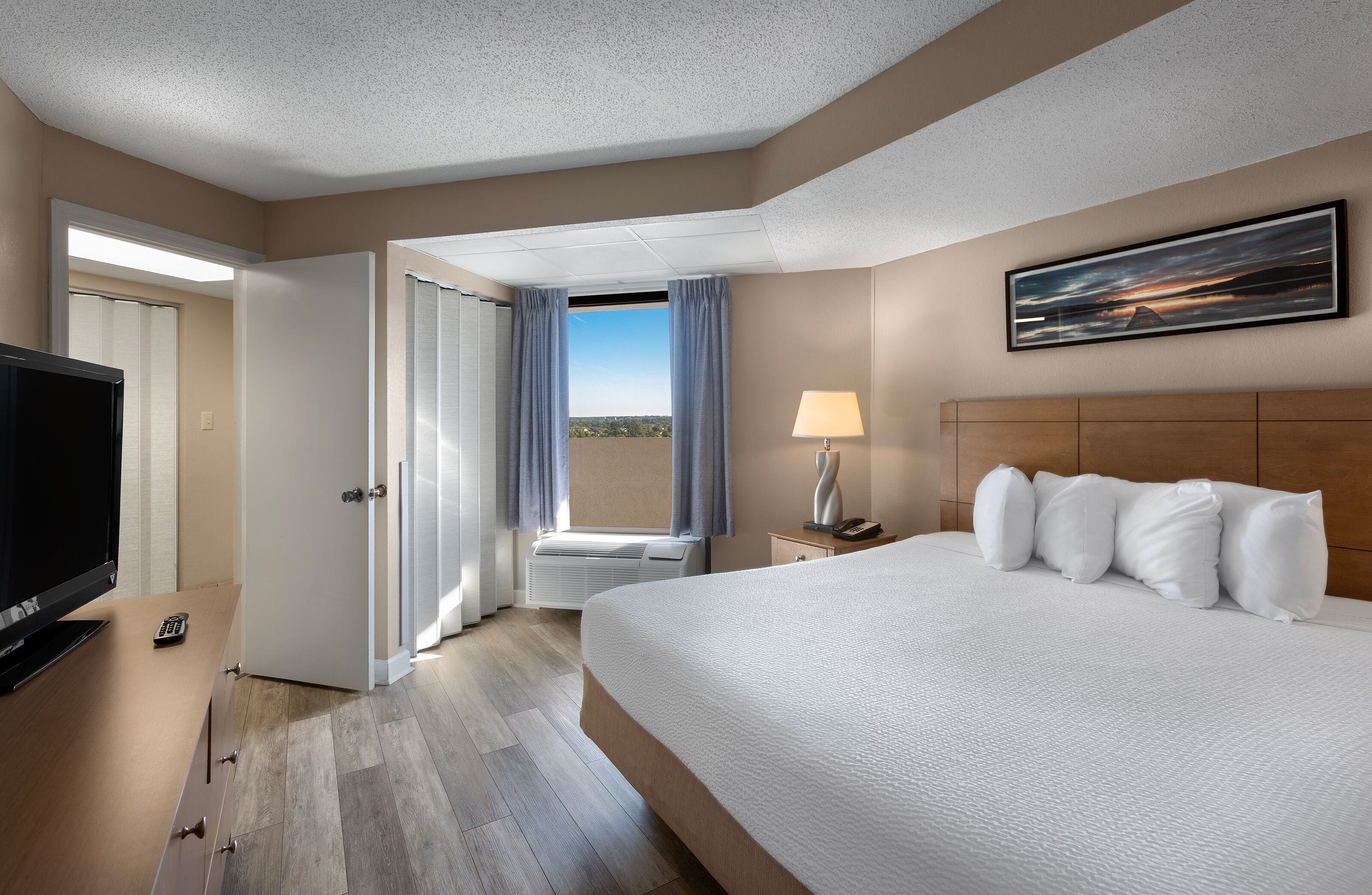 A hotel room with a large bed, a TV, a desk with a remote, a lamp, and a painting on the wall.