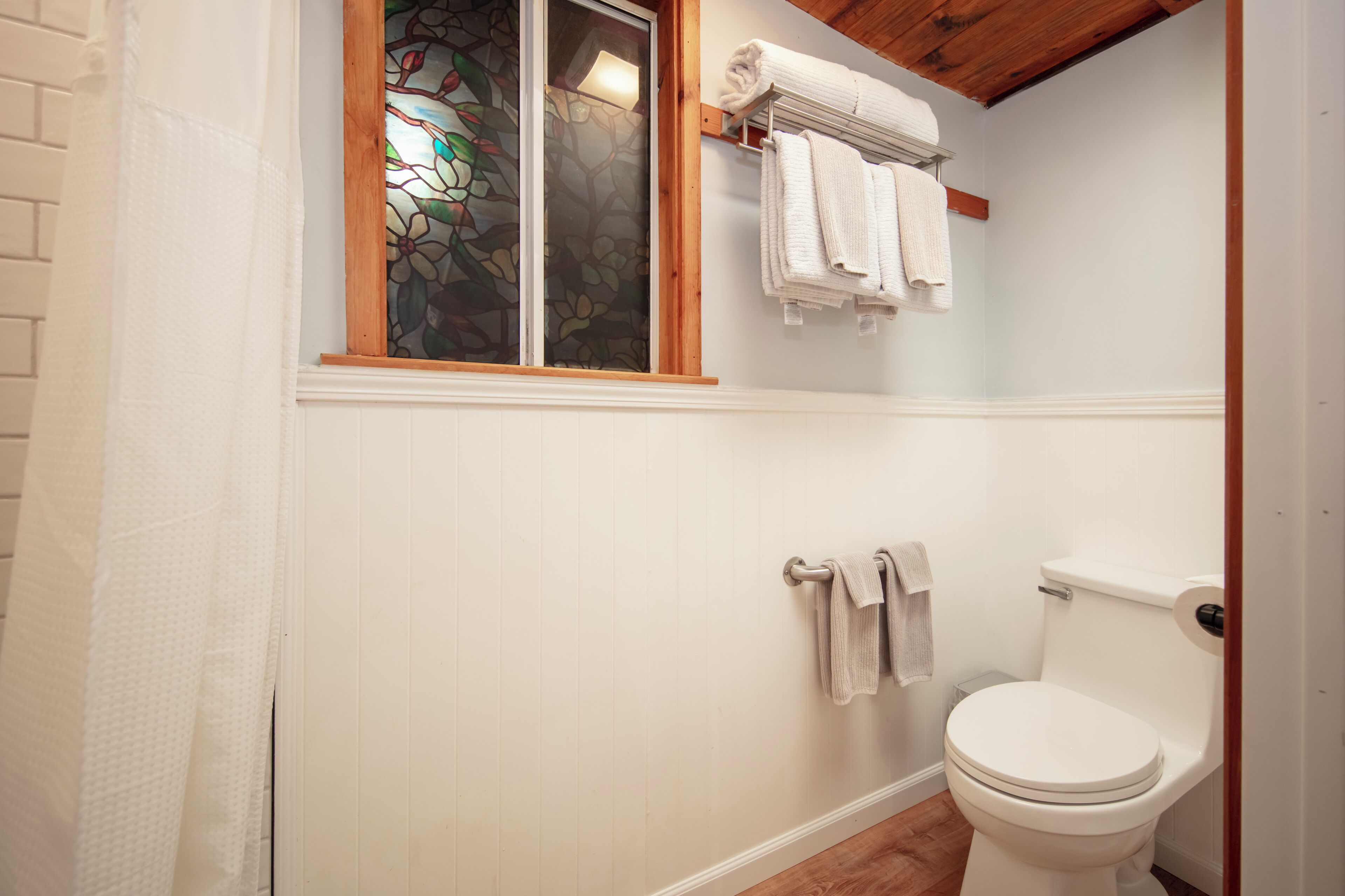 Ein Badezimmer mit einer weißen Toilette, einem Fenster mit Holzrahmen und farbigem Glas sowie einem Handtuchhalter mit gefalteten Handtüchern.