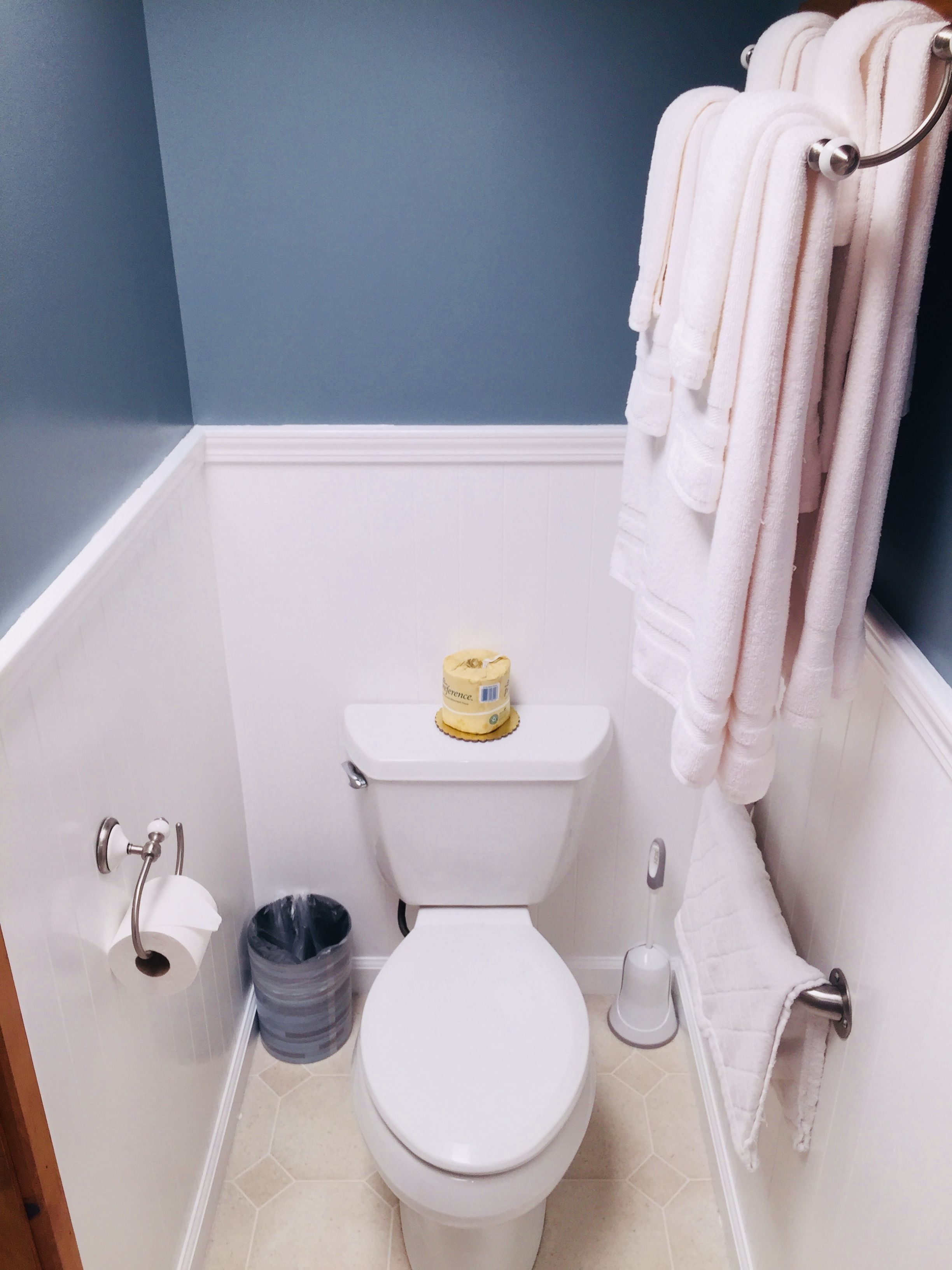 Un baño con inodoro, rollo de papel higiénico, un cubo de basura y un perchero con toallas.