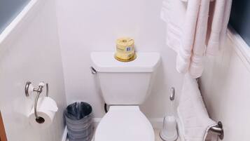 Un baño con inodoro, rollo de papel higiénico, un cubo de basura y un perchero con toallas.