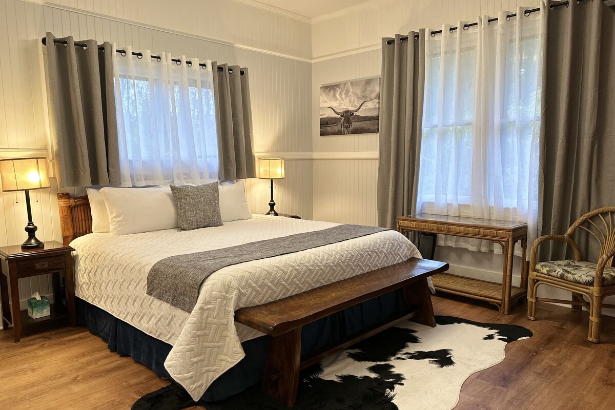 A neatly arranged bedroom with a large bed, bedside tables, a wooden bench, a cowhide rug, and a wicker chair.