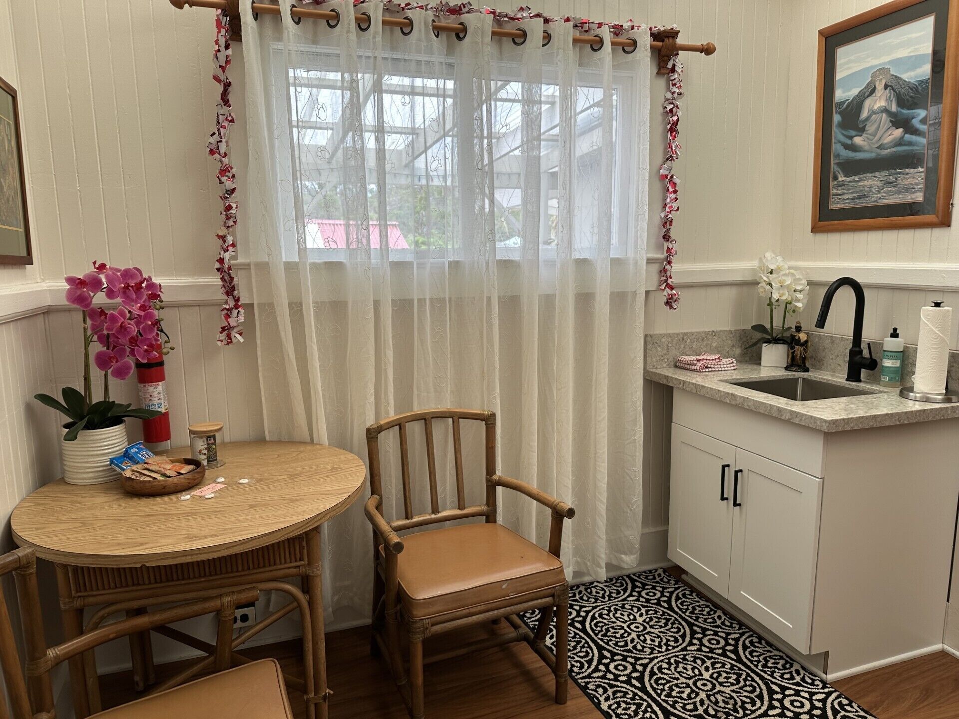 Una cocina pequeña con un lavamanos, una mesa de comedor con sillas y una ventana con cortinas traslúcidas.