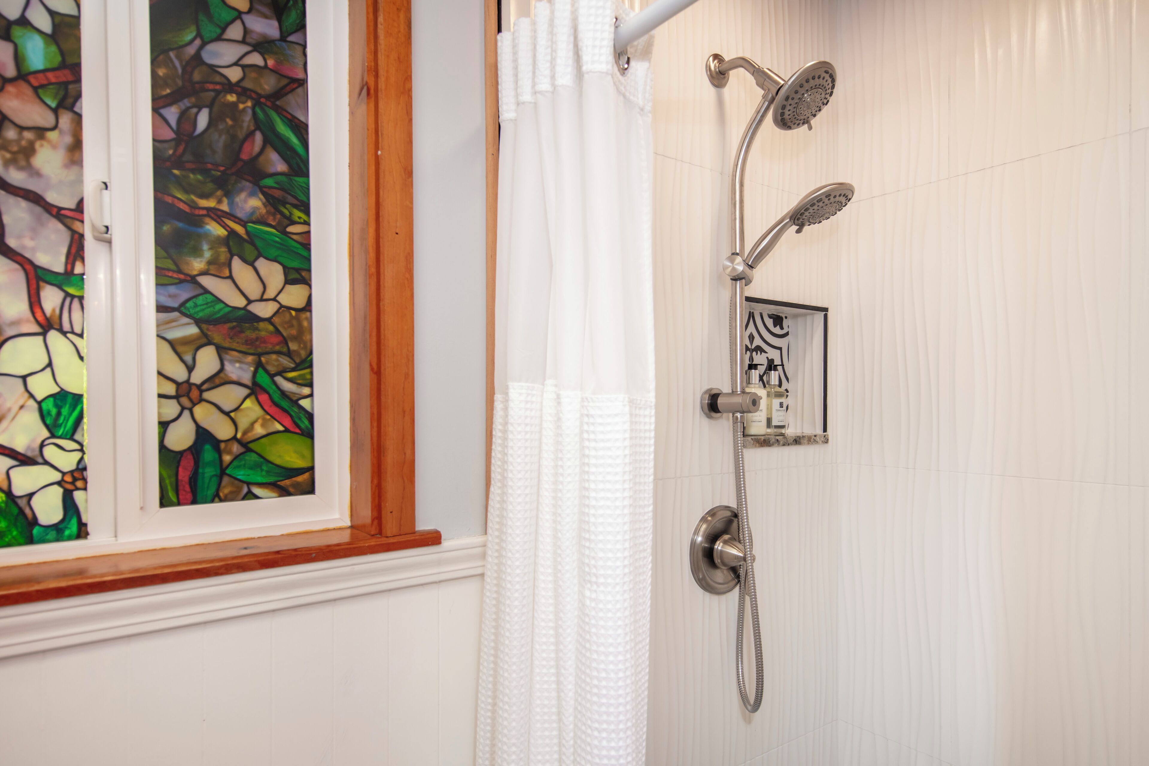 Un baño con ducha, una ventana con vidrieras y una pared de azulejos blancos.