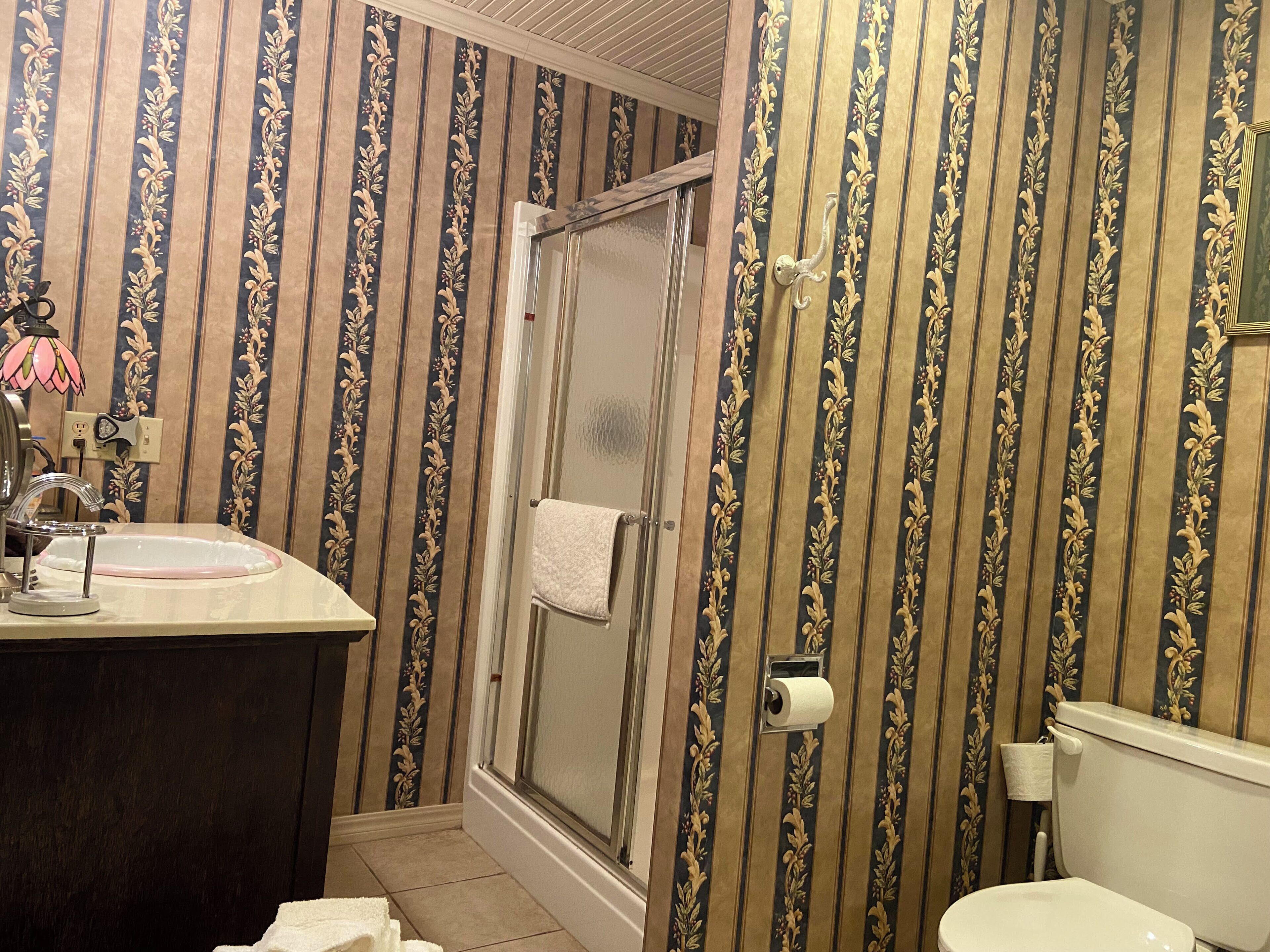A bathroom with striped wallpaper, a sink with a pink faucet, a shower with a glass door, and a toilet.